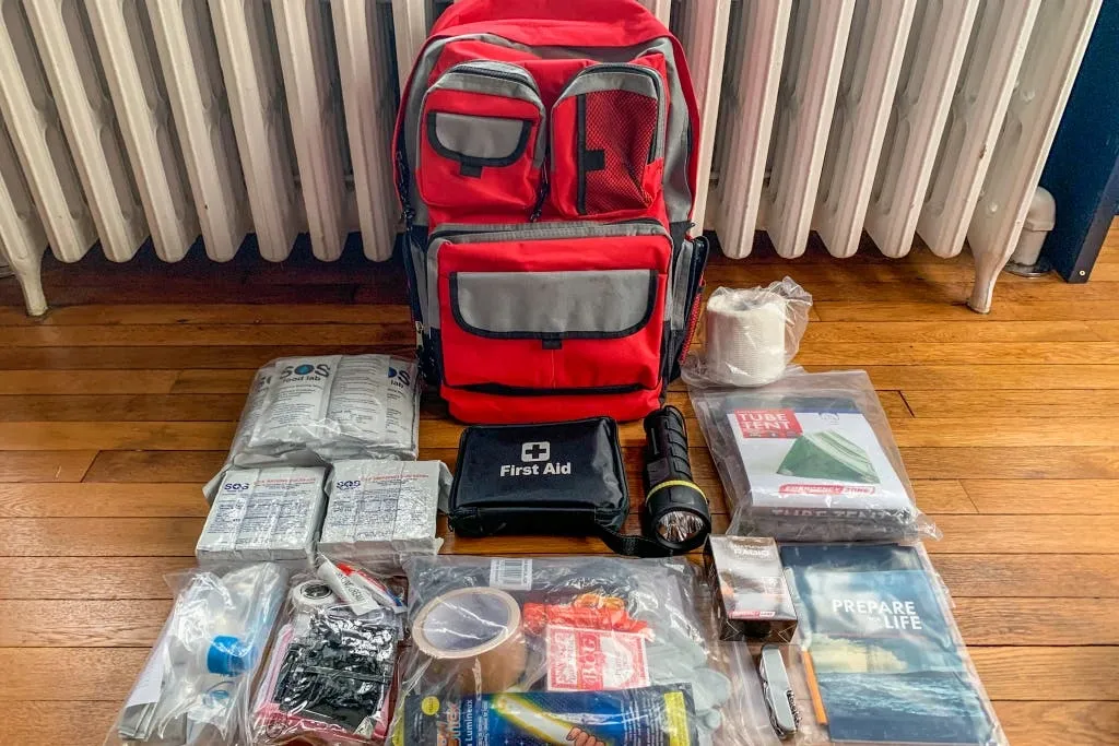 Red go bag surrounded by emergency supplies including first aid kit, flashlight, water packets, and survival items on wooden floor.