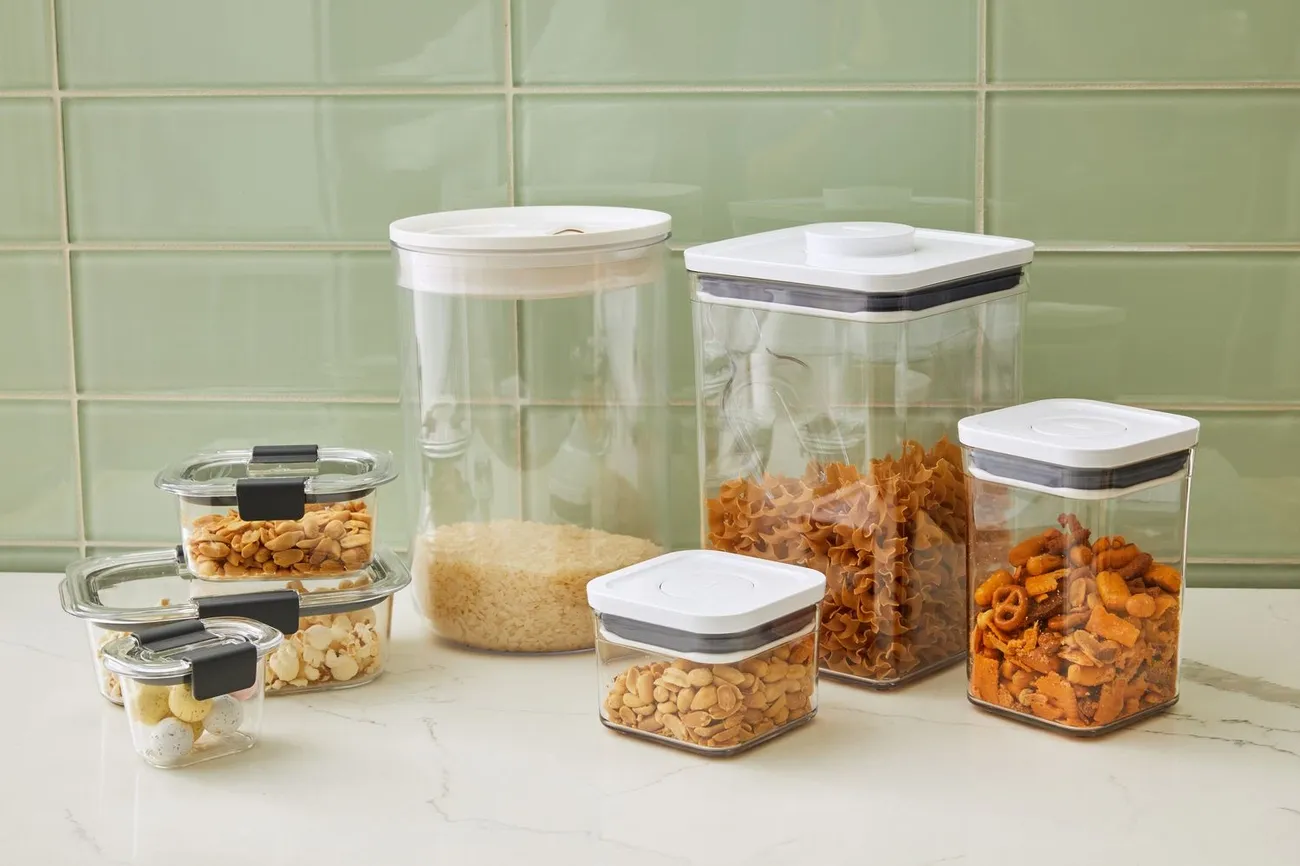 Various clear food storage containers with white and black lids filled with snacks and dry goods on a countertop
